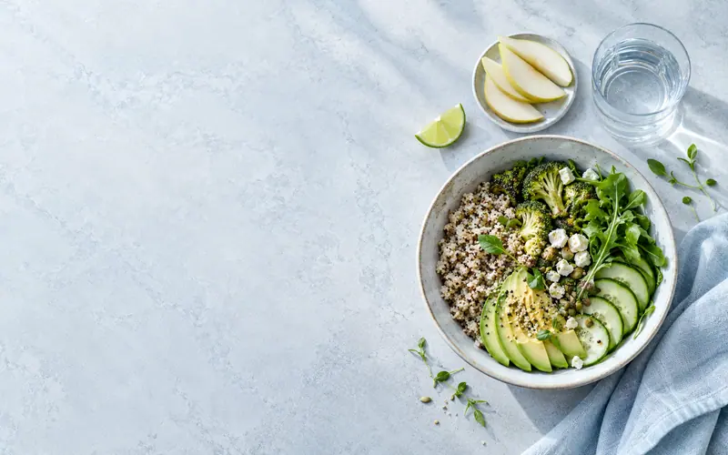 A bowl of quinoa, avocado, broccoli and cucumber on a pale marble surface with a glass of water and a bowl of sliced pear.